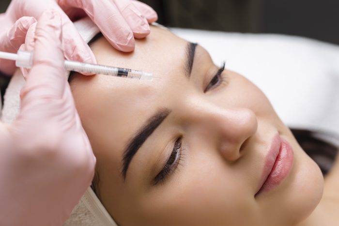 Close up of hands of young cosmetologist injecting botox in female face. She is standing and smiling. The woman is closed her eyes with relaxation