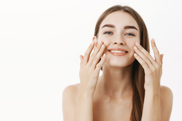 Waist up shot of charming relaxed and gentle young woman making cosmetological procedure applying facial cream on face with fingers and smiling broadly feeling perfect, taking care of skin