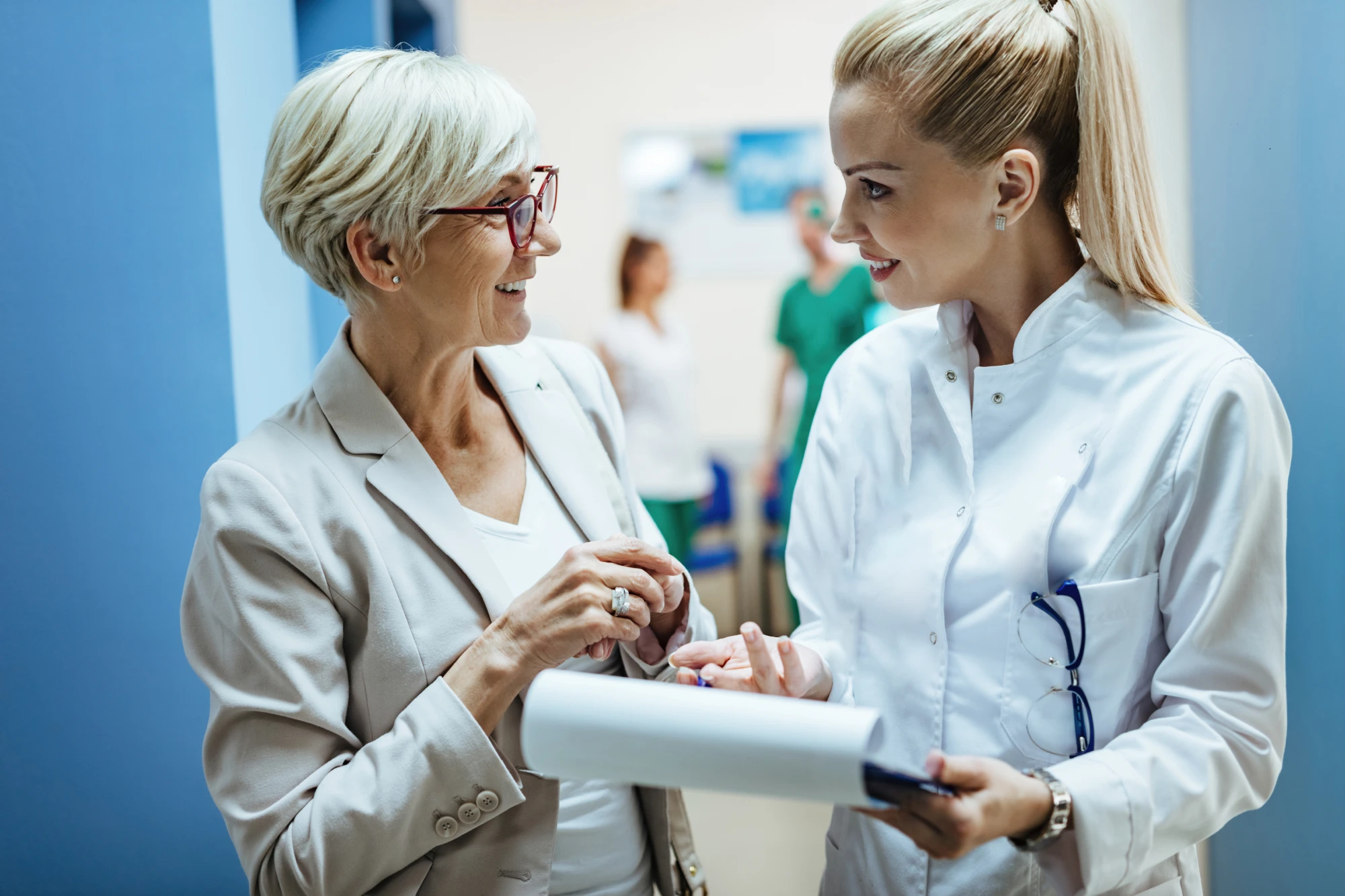 Two women engaged in a conversation in a medical setting. One is an older woman with short white hair and glasses, wearing a beige blazer. The other is younger with long blonde hair, dressed in a white medical coat and holding a clipboard. They are smiling warmly.