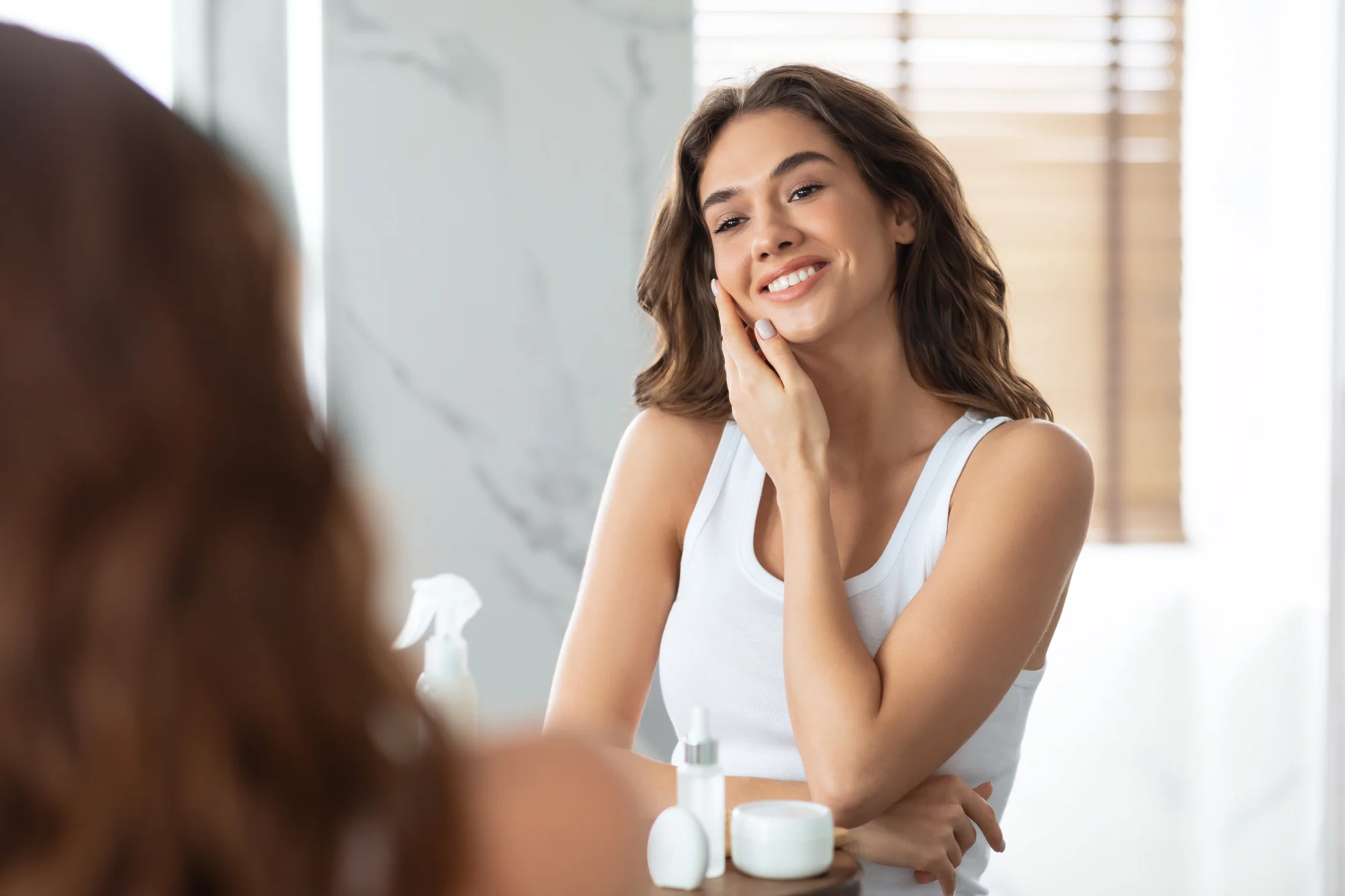 A woman with long brown hair and wearing a white tank top is smiling and touching her face while looking into a mirror. Skincare products are visible on the counter in front of her. The setting appears to be a bathroom with a blurred background.