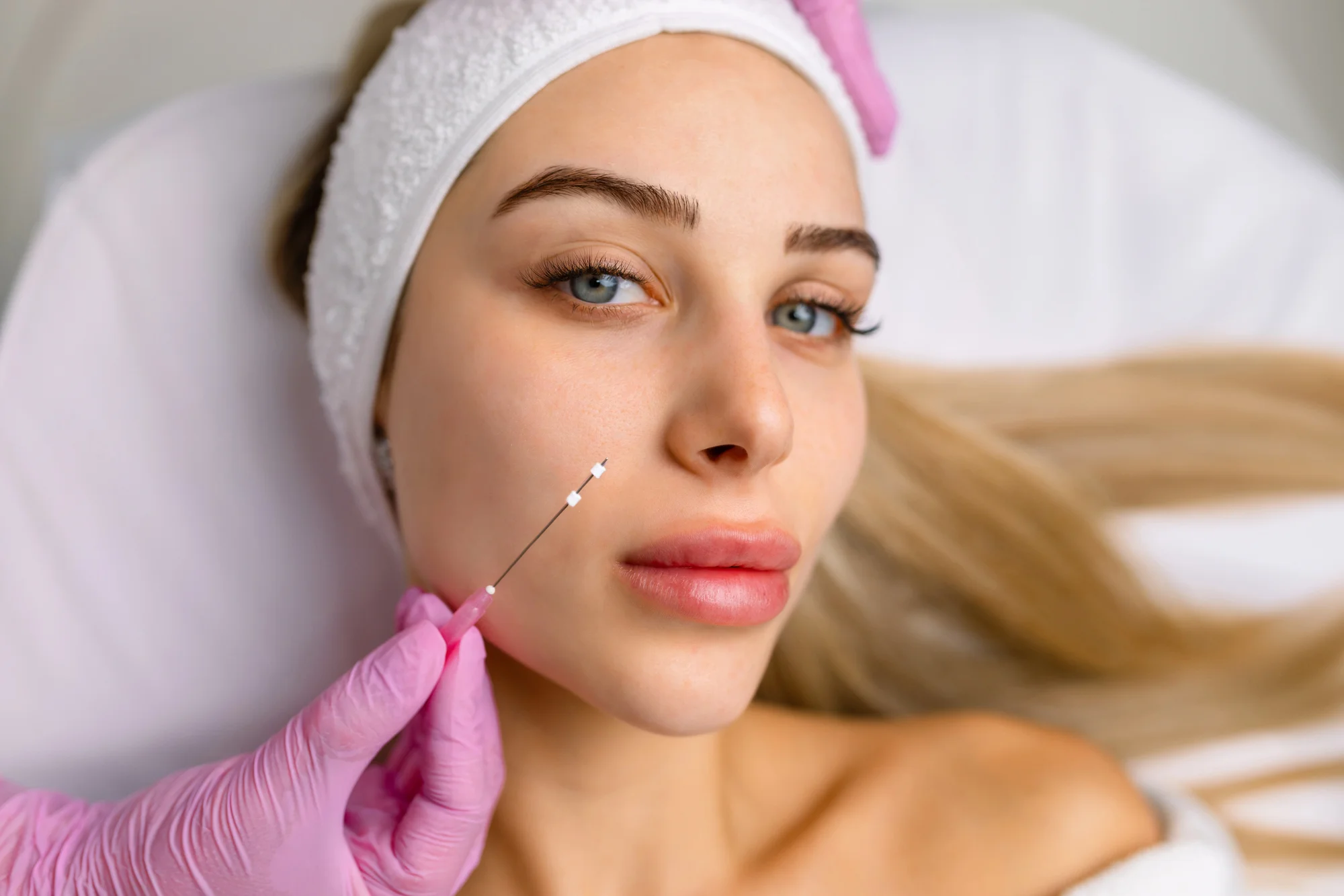 A woman with long blonde hair lies on a white bed, wearing a white headband and a pink glove. A close-up view shows a needle near her cheek, hinting at a cosmetic procedure involving threads. She gazes at the camera with a calm expression.