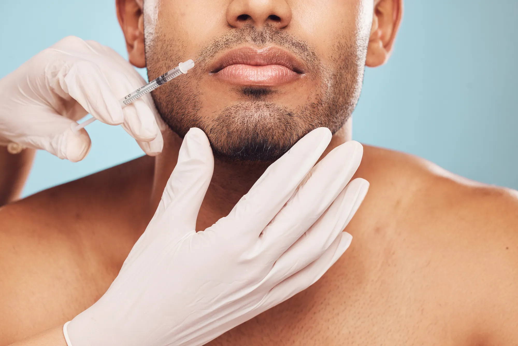 A person receiving a cosmetic injection in the lower face. They are wearing no shirt, and their facial hair is trimmed. Hands in white gloves are administering the injection, with a blue background.