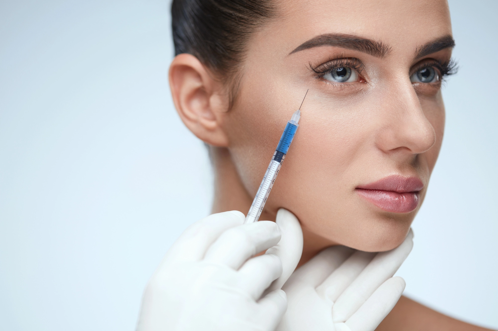 A close-up of a woman receiving a cosmetic injection in under eye area. She has light makeup, and her hair is pulled back. A gloved hand holds a syringe with a needle poised at her temple. The background is softly blurred.