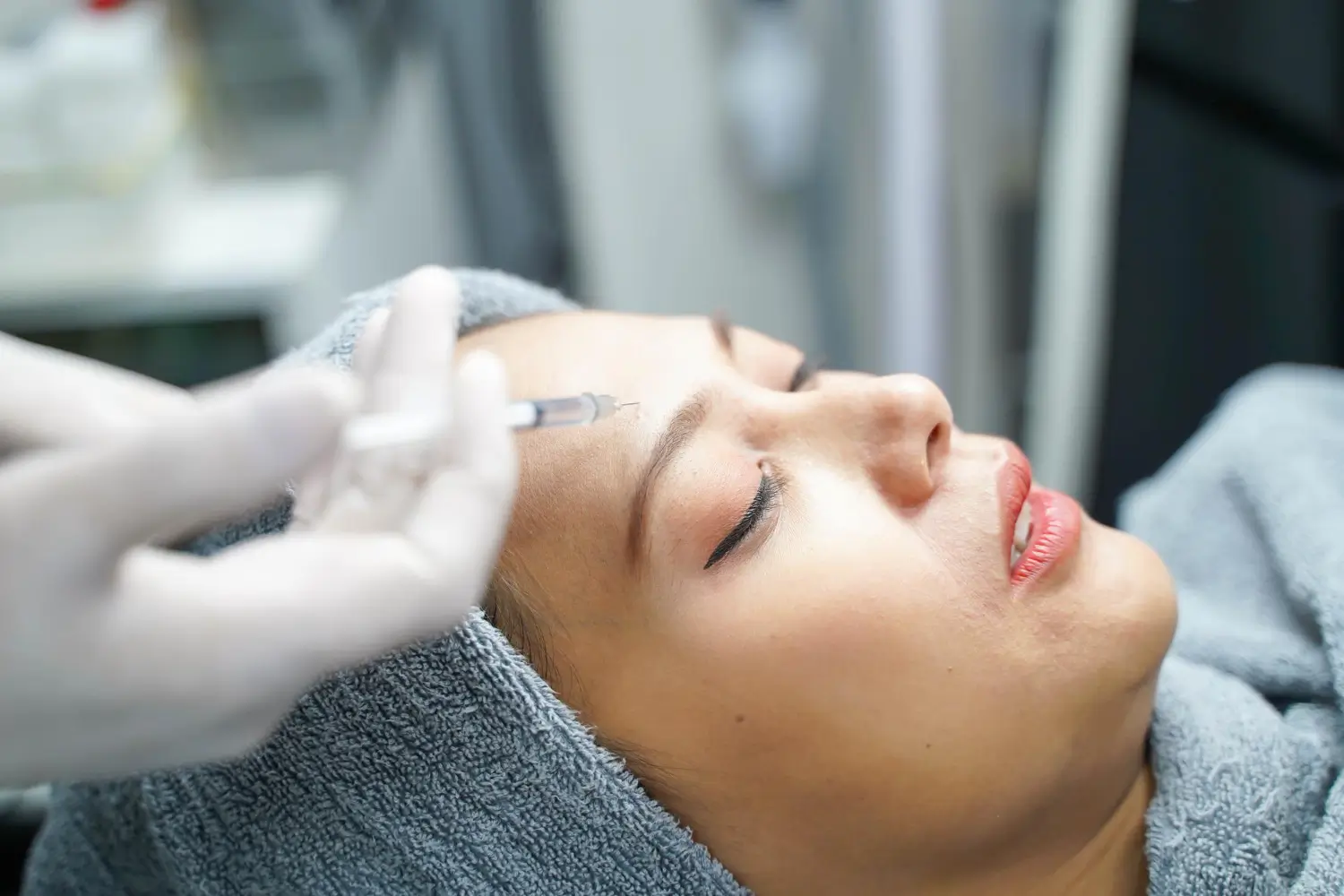 A person receives a cosmetic injection in the forehead. They are lying down, covered with a gray towel, while a gloved medical professional administers the injection. The setting appears to be a clinic or spa.