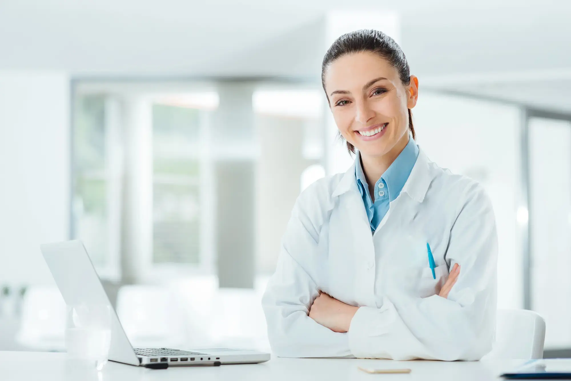 A smiling woman in a white lab coat sits at a desk with a laptop in a bright, modern office. She is crossing her arms and has a pen in her breast pocket. The setting conveys a professional and clean environment.