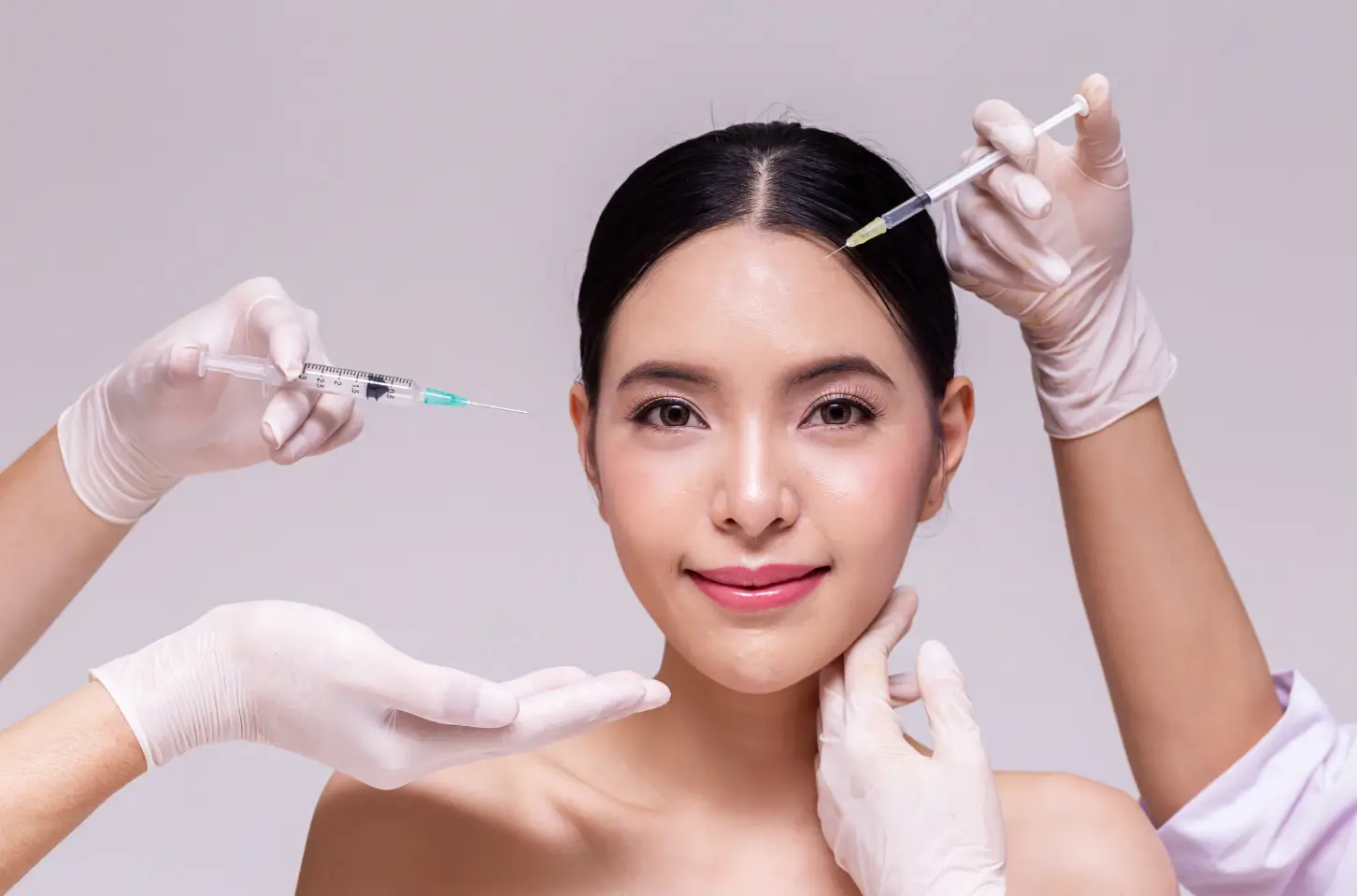A confident woman receives a cosmetic injection in her forehead while two other hands in white gloves frame her face.